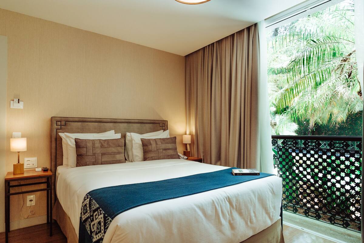 A hotel room with a large bed, a nightstand with a lamp, a window with curtains, and a view of greenery outside.