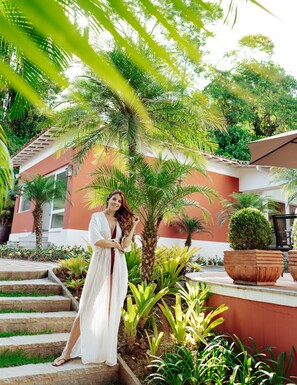 A woman standing on stone steps in a lush garden with palm trees and a red building in the background.