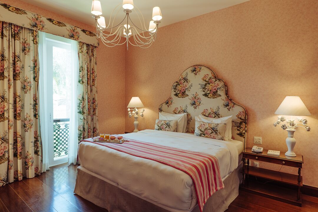 A hotel room with a large bed, floral headboard, two bedside lamps, a nightstand, and a view of the outdoors through a door with curtains.