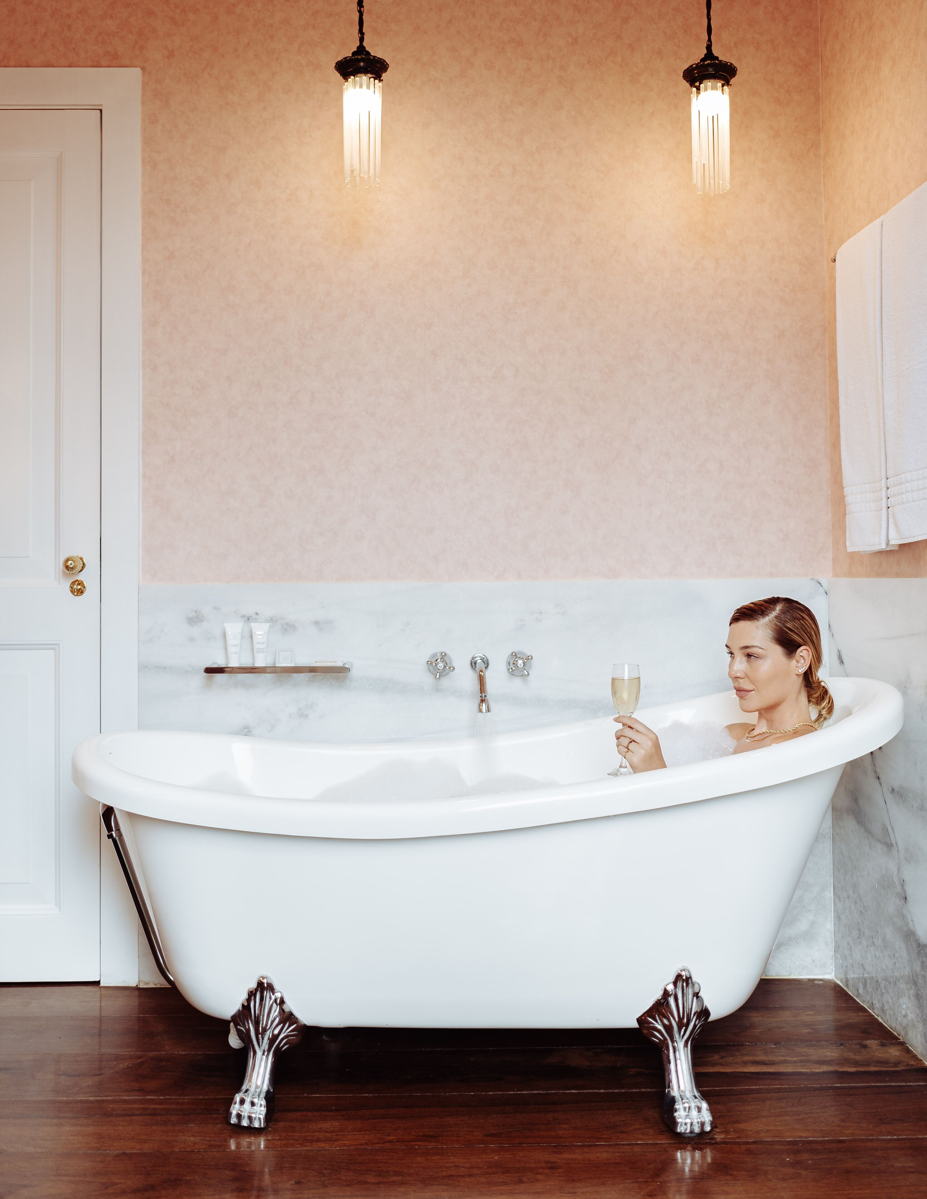 Un baño con una bañera independiente, dos lámparas colgantes, una pared de mármol y suelo de madera.