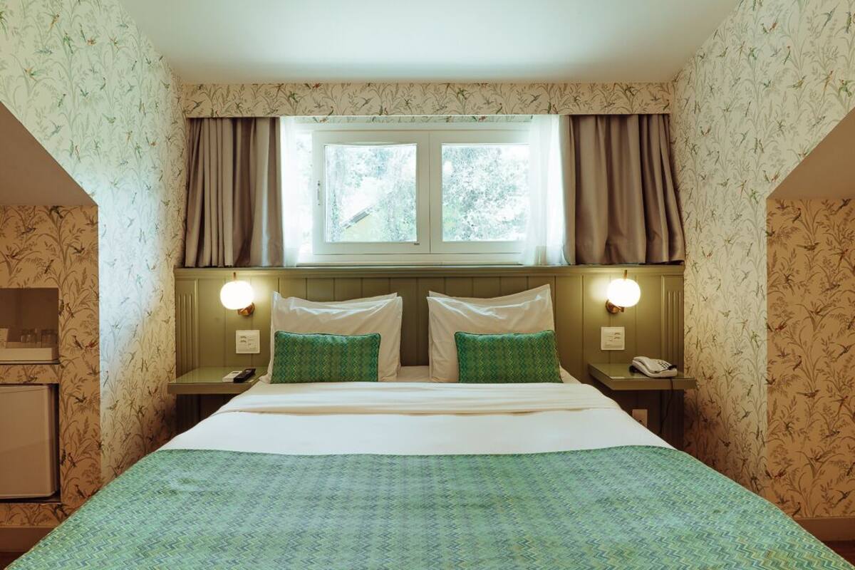A hotel room with a bed, two bedside tables, a nightstand, a window with curtains, and floral wallpaper.