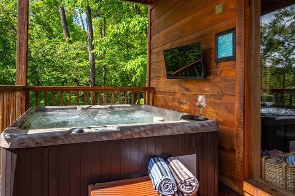 A wooden cabin with a hot tub, a wooden deck, and a view of a forest.
