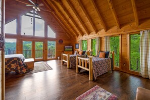 A cabin with a high ceiling, wooden beams, a large bed, two smaller beds, a TV, and large windows offering a view of the forest.