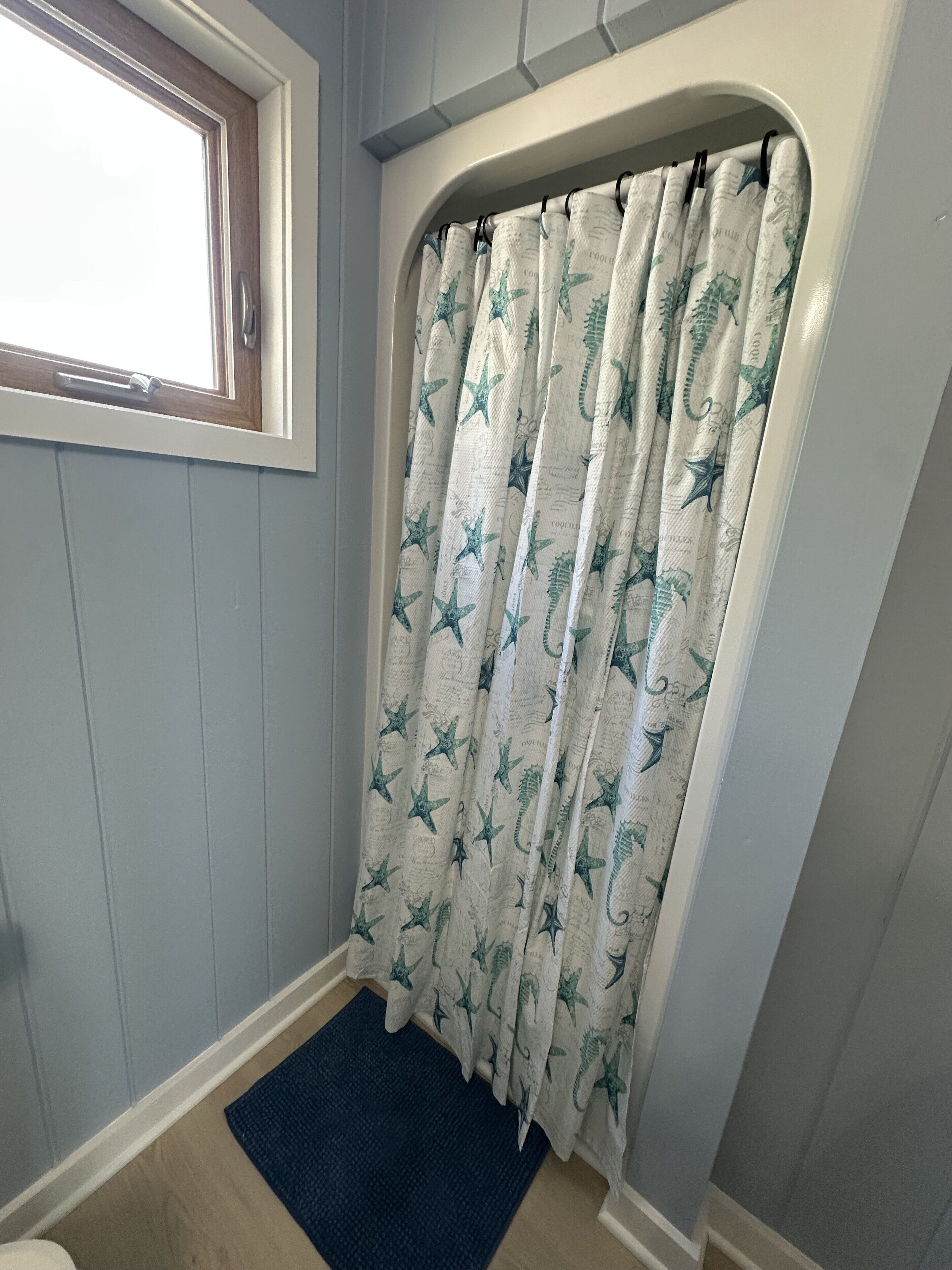 A bathroom with a shower curtain featuring a starfish and sea creature pattern, a window, and a blue rug.