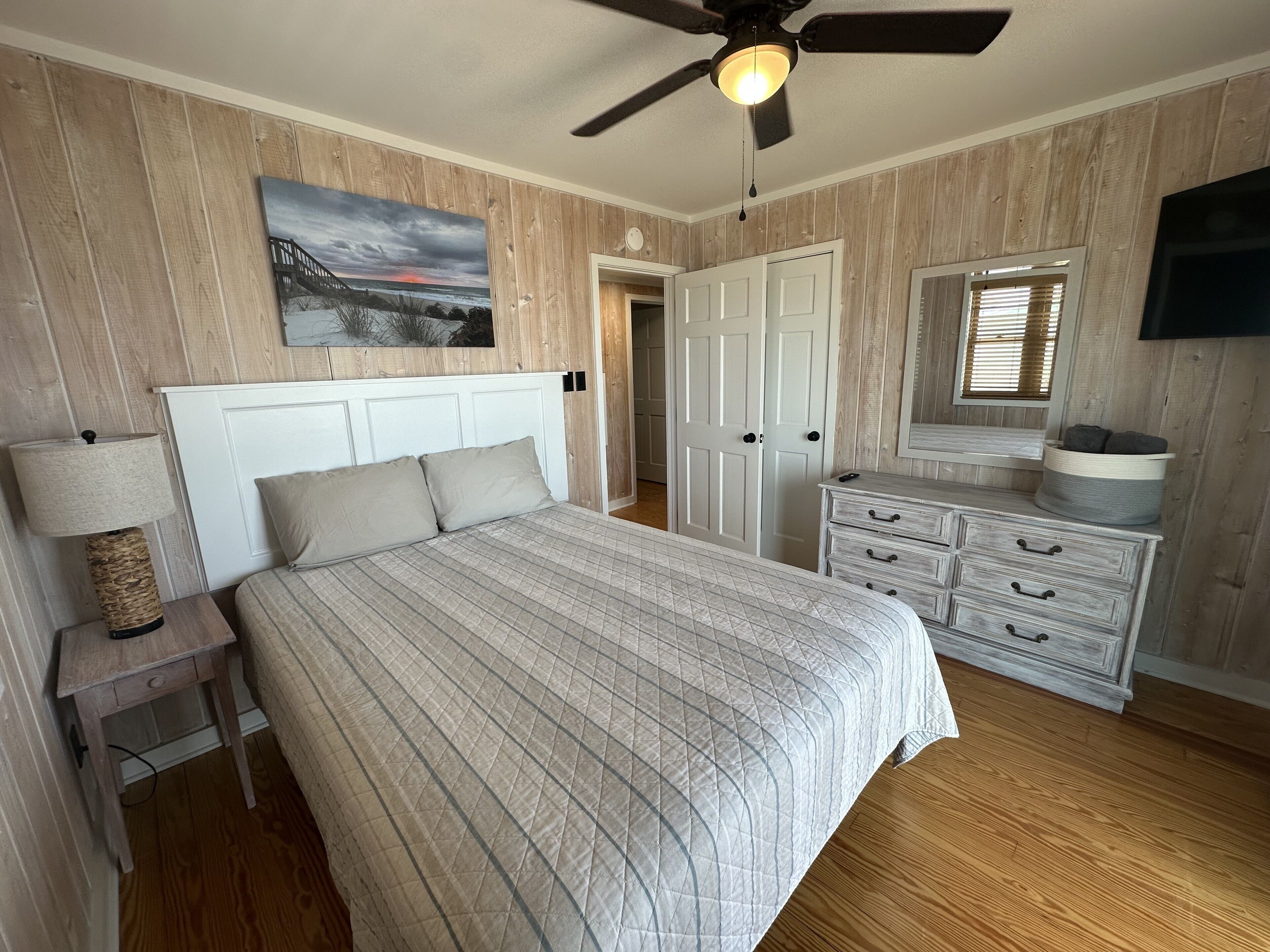 Une chambre à coucher comprenant un lit, une table de chevet, une commode, un ventilateur de plafond, un tableau et une porte.