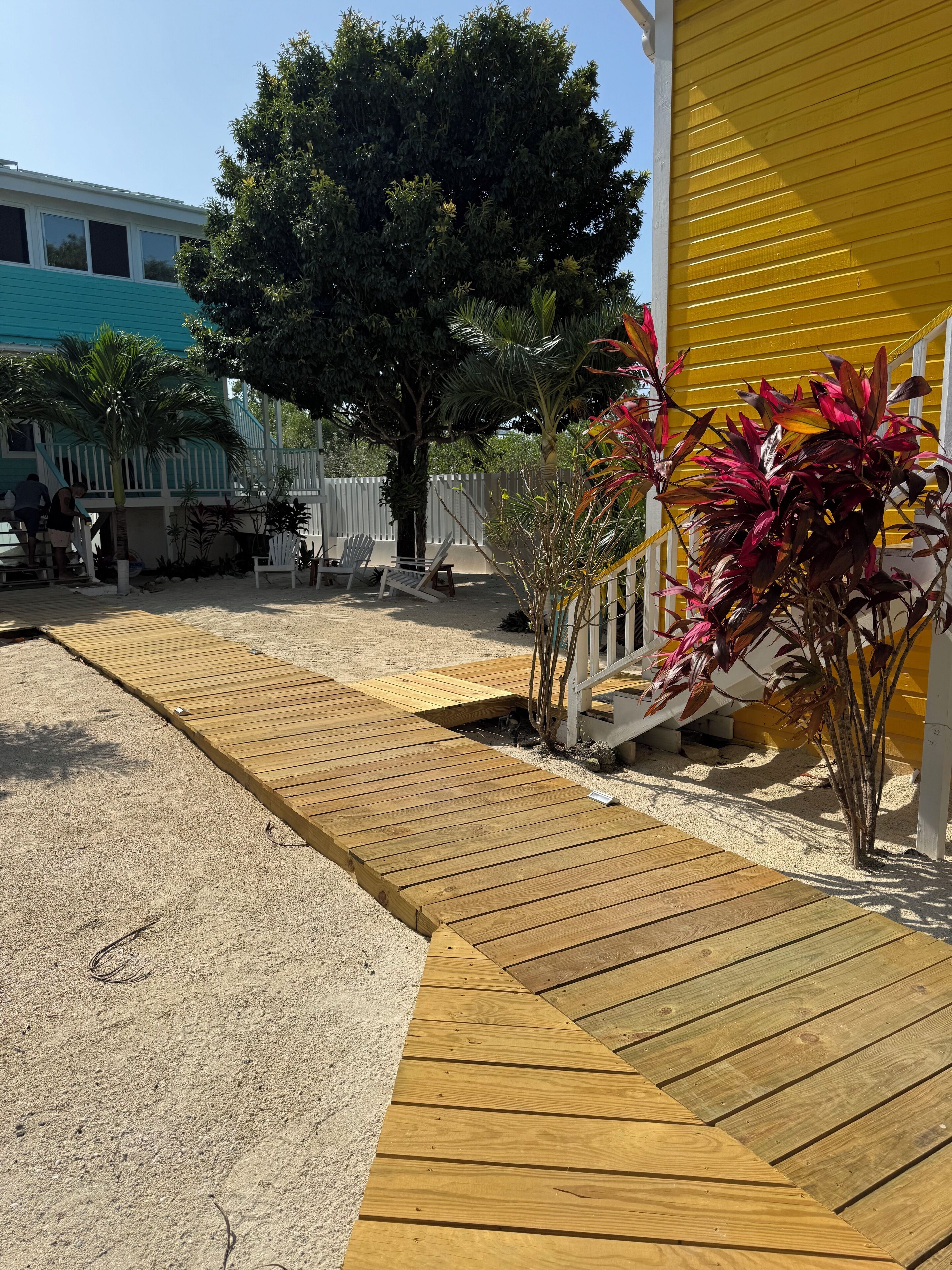 Una terraza de madera que conduce a una casa de color amarillo, rodeada por una valla blanca y diversas plantas.