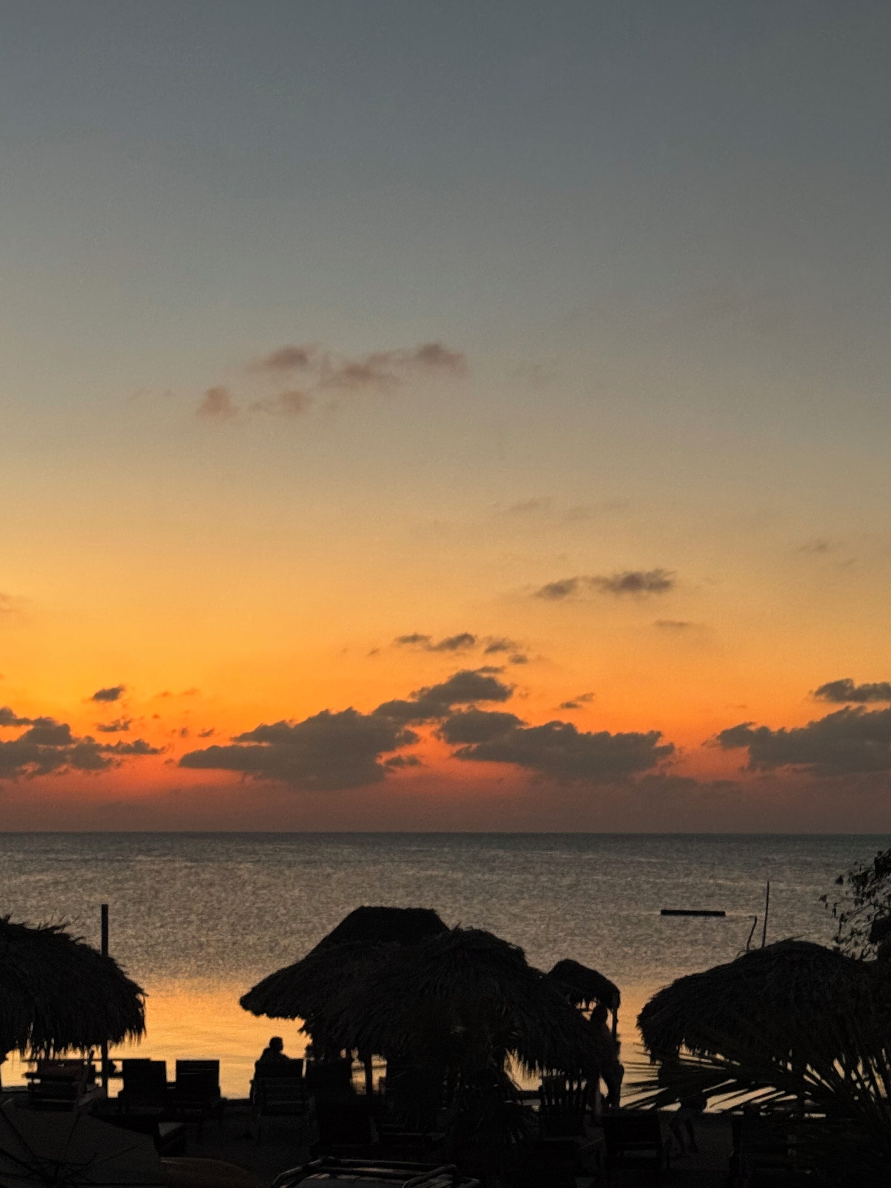 Escena de playa con sombrillas de paja y sillas reclinables, un mar tranquilo y un cielo al atardecer.