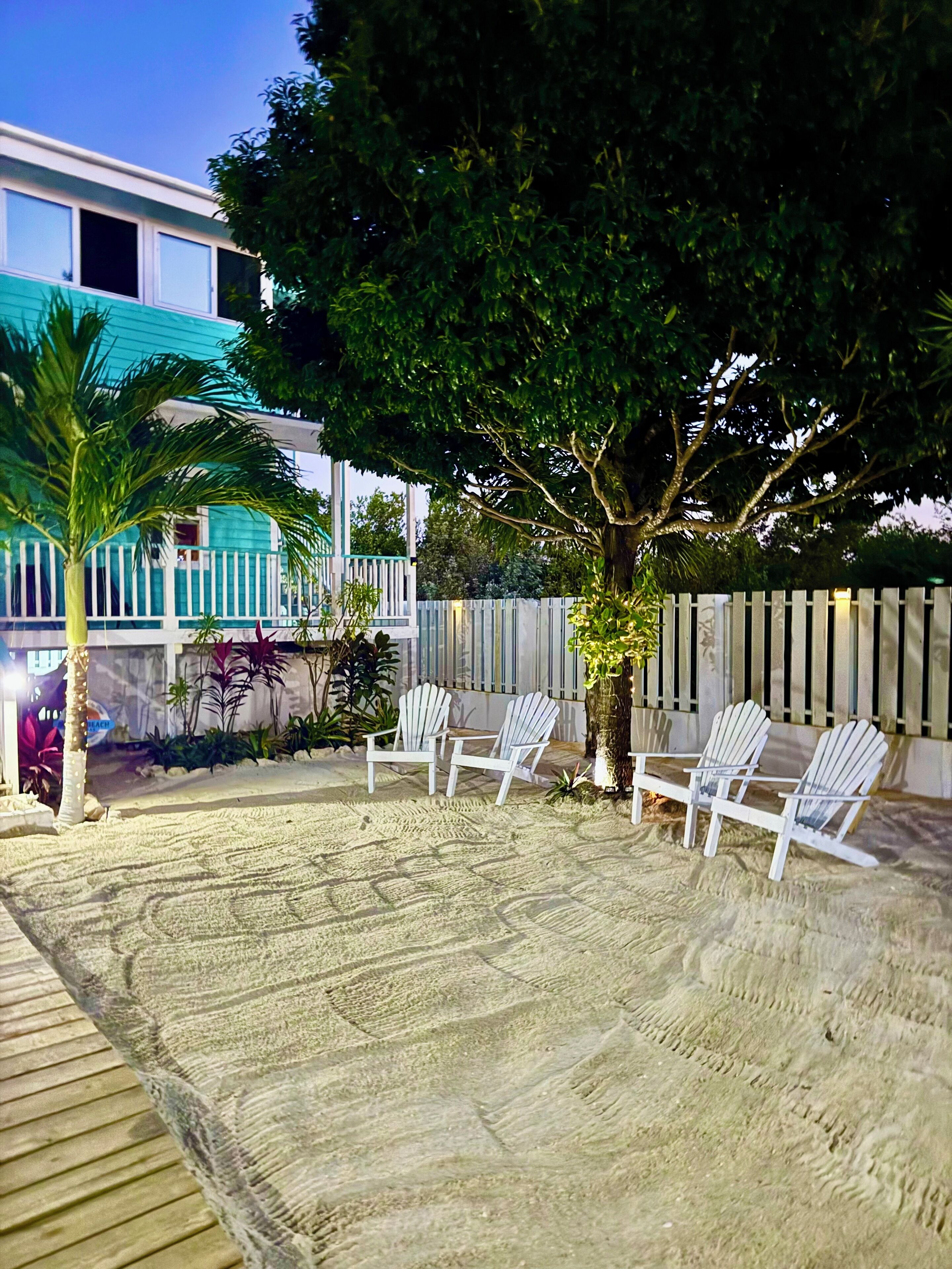 A sandy outdoor area with white Adirondack chairs, a palm tree, and a wooden deck.
