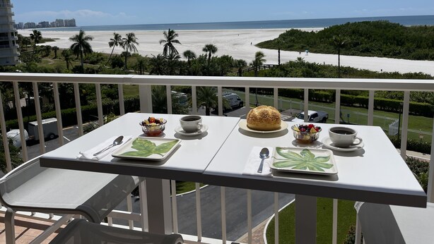 A balcony with a table set for two, overlooking a beach and ocean.