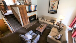A living room with a fireplace, sofa, and coffee table. There is a TV on a wooden shelf above the fireplace. - Beautiful Ski In/Out Cedars Townhome with Private Hot Tub (Breckenridge)