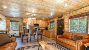 L’intérieur chaleureux d’un chalet avec un plafond en bois, un coin cuisine, un espace salon avec un canapé et des fauteuils, et de grandes fenêtres offrant une vue sur l’extérieur.