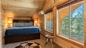 Une chambre dans un chalet en bois, avec un lit, deux lampes de chevet, une fenêtre donnant sur la forêt et une décoration rustique comprenant un crâne accroché au mur.