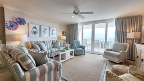 A spacious living room with a striped sofa, a blue armchair, and a white coffee table. There are decorative glass pieces on the wall and a ceiling fan. - Hamilton 202, Oceanfront, Litchfield by the Sea (Pawleys Island)