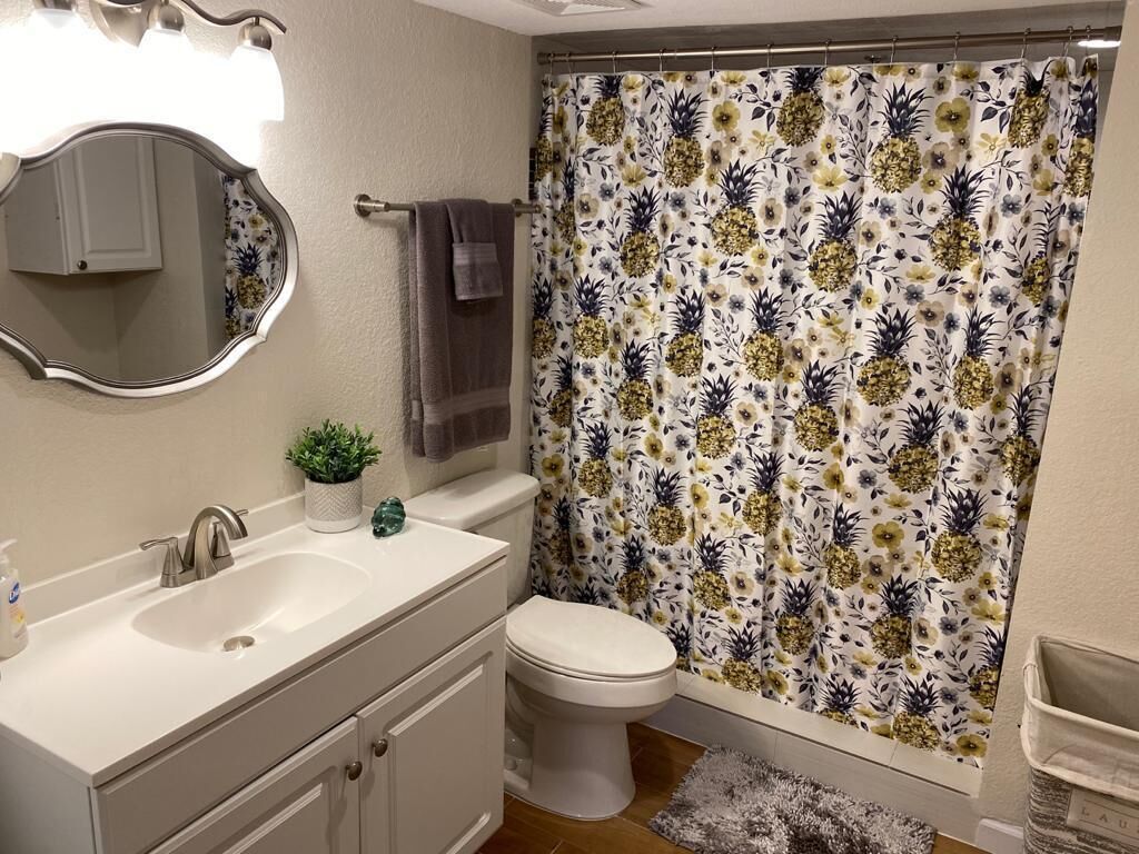Een badkamer met een witte wastafel, toilet, douchegordijn met bloemendesign, spiegel en een kleine plant op de wastafel.