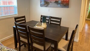 A dining area with a wooden table and chairs, a painting on the wall, and a window with blinds. - The Pink House- minutes walk to beach!! (Galveston)