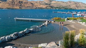 A lakeside scene with a dock extending into the water, surrounded by rocks and a grassy area with a few people walking. - Welcome to Summersalts at Lake Chelan (Chelan)
