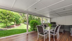 Un espace repas extérieur moderne, doté d’une table et de chaises en verre, d’une terrasse en bois et offrant une vue sur un jardin luxuriant.