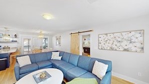 A modern living room with a blue sectional sofa, a white coffee table, and a kitchen area with a dining table. - Daydream Oasis with private pool, screen porch with dining, sleeps 8! (Isle of Palms)