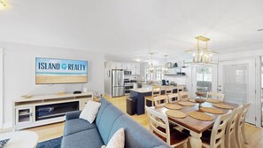 A modern kitchen with stainless steel appliances, a dining area with a wooden table and chairs, and a living room with a blue sofa and a flat-screen TV displaying 'ISLAND REALTY'.
