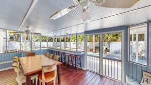 A covered outdoor dining area with a wooden table and chairs, a ceiling fan, and large windows.