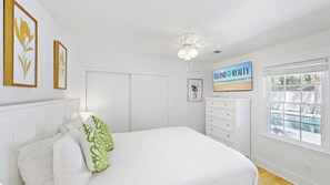 Une chambre avec un lit, une commode, un ventilateur de plafond, une fenêtre donnant sur une piscine et des œuvres d’art encadrées accrochées au mur.