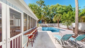 A pool area with a wooden deck, lounge chairs, and a view of a house with large windows and a blue exterior.