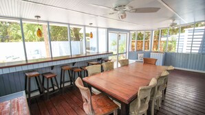 Un espace intérieur spacieux avec une grande table en bois et plusieurs chaises, de grandes fenêtres donnant sur un paysage verdoyant et un ventilateur de plafond.