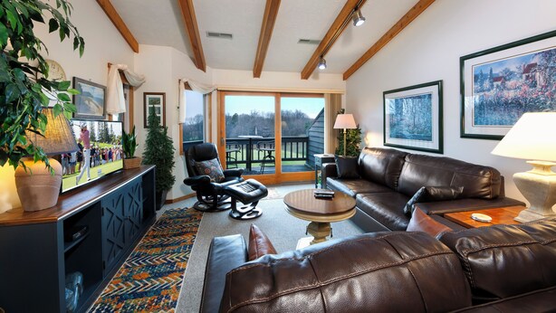 A cozy living room with leather furniture, a flat-screen TV, and a large window offering a view of the outdoors. - Balcony with Lake Michigan and golf view (Cedar)