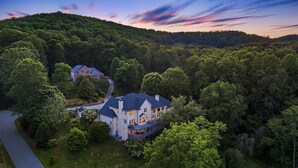 A large, two-story house with a dark roof, surrounded by dense forest and a river. - Luxury Estate w/ Hot Tub & Game Room + Biltmore Pass | 18 Min to Asheville (Asheville)