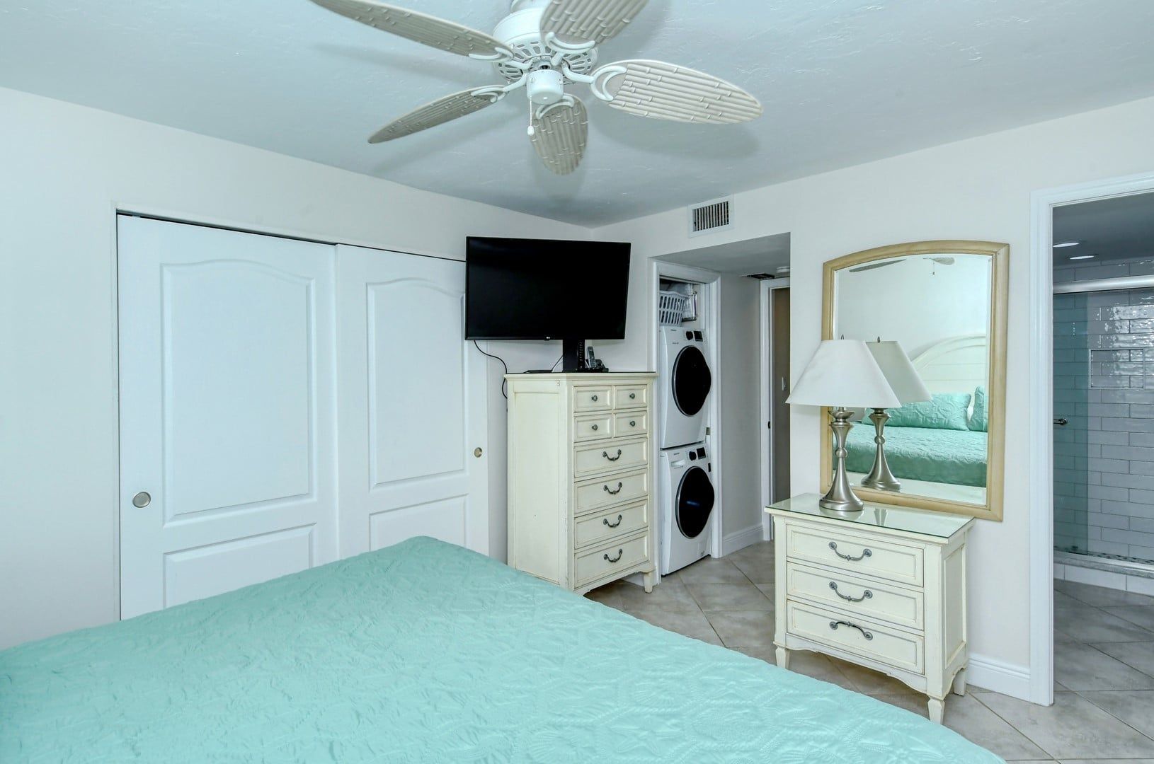 Een slaapkamer met een bed, een plafondventilator, een televisie, een wasmachine, een dressoir en een spiegel.