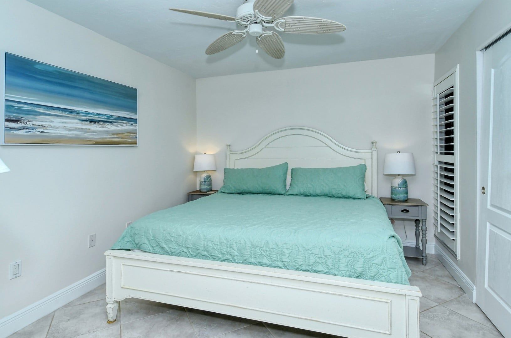 A bedroom with a bed, two bedside tables, a ceiling fan, a painting on the wall, and a door with blinds.