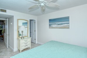 A bedroom with a bed, a dresser with a mirror, a ceiling fan, and a beach-themed painting. - Firethorn 210- Beautiful gulf side, ground-level 2BR/2BA unit! (Sarasota)
