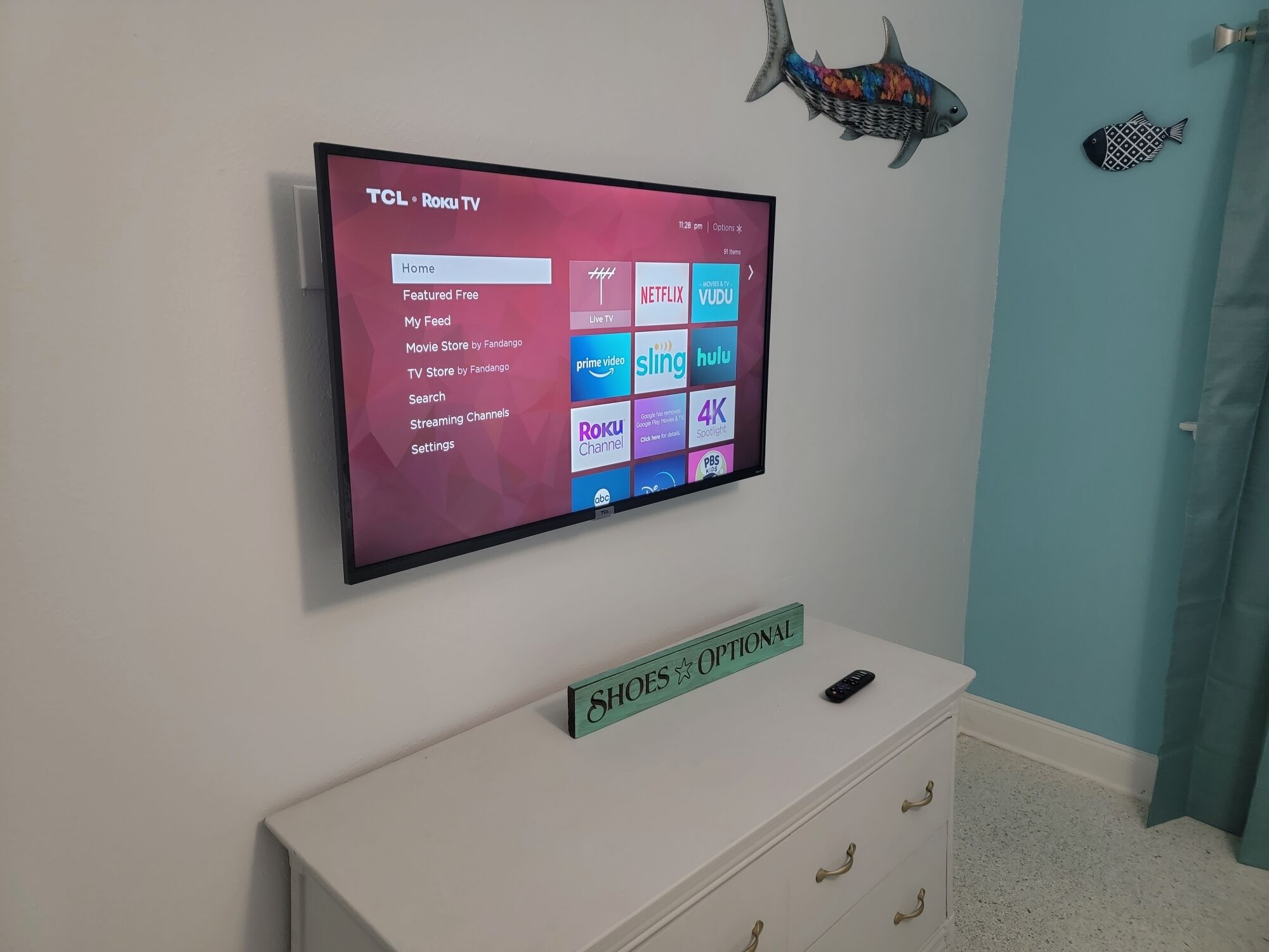 A TCL Roku TV mounted on a wall with a white dresser below it.