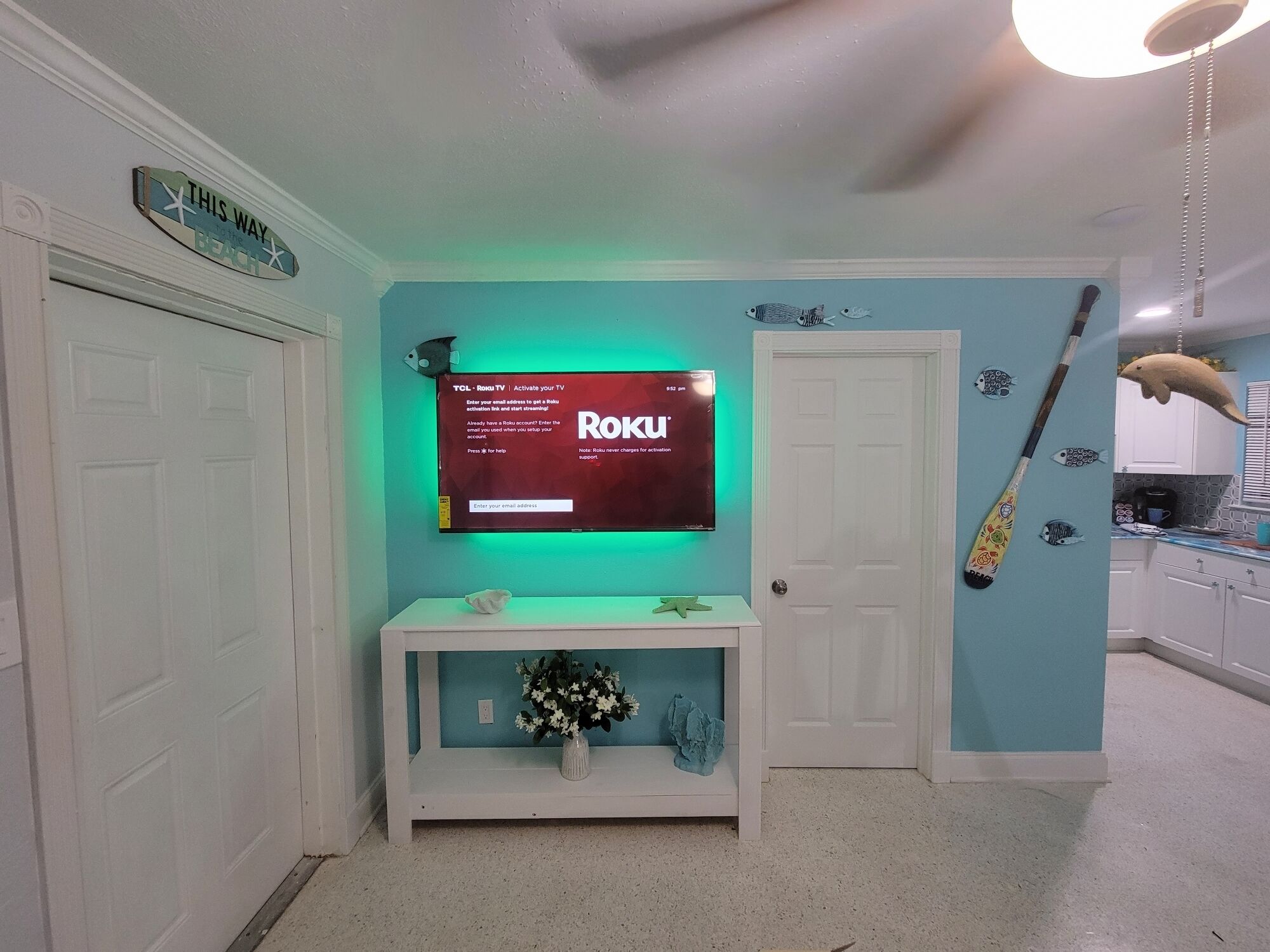 Una habitación con una televisión montada mostrando la pantalla de activación de Roku, una consola blanca con un florero y una puerta con un letrero que dice 'Por aquí'.