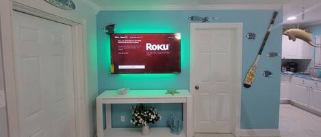 Una habitación con una televisión montada mostrando la pantalla de activación de Roku, una consola blanca con un florero y una puerta con un letrero que dice 'Por aquí'.
