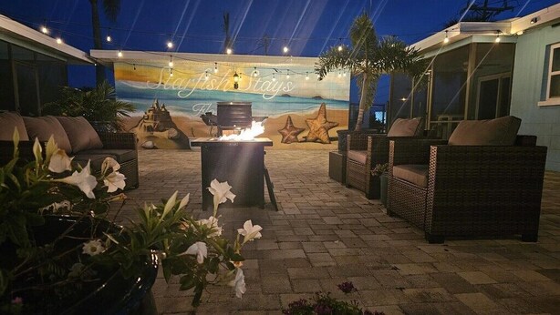 Eine Terrasse im Freien mit Korbmöbeln, einer Feuerstelle und einem Wandbild mit Strandmotiv.