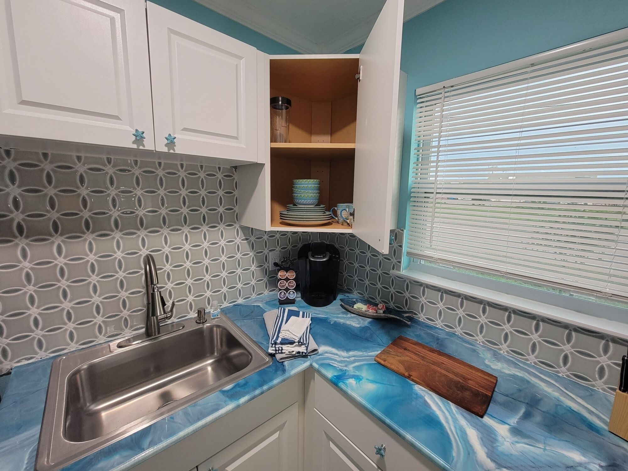 A kitchen with a blue marble countertop, a stainless steel sink, white cabinets, and a window with blinds.