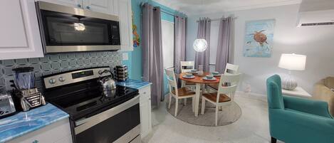 Une cuisine compacte équipée d’un four à micro-ondes, d’une cuisinière et d’un mixeur, un coin repas avec une table et des chaises, et un fauteuil de couleur turquoise.