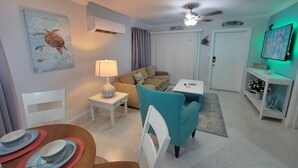 Ein Wohnzimmer mit einer Couch, einem türkisblauen Sessel, einem Couchtisch, einer Lampe und einem Esstisch mit Tellern.