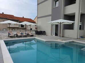 Piscina com espreguiçadeiras e guarda-sóis, cercada por um edifício com telhado em azulejos.
