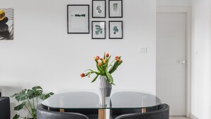 A dining area with a round glass table, two black chairs with wooden legs, a white vase with flowers, and a hanging light fixture.
