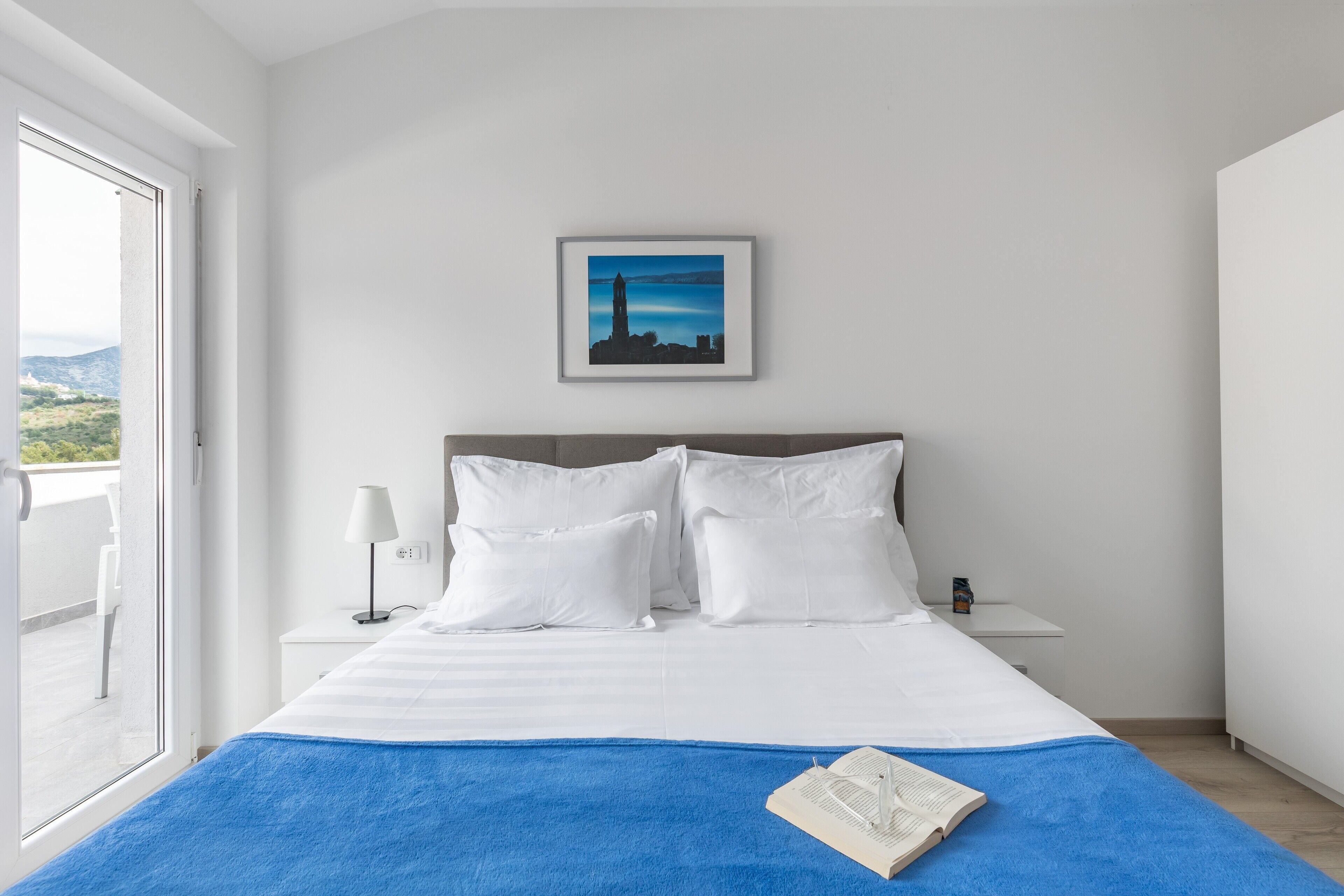 A bedroom with a bed, white pillows, a blue blanket, a framed picture on the wall, a bedside table with a lamp, and a window with a view of the outdoors.