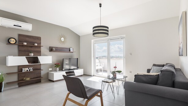 A modern living room with a grey sofa, a TV, a dining table with chairs, and a large window offering a city view. - Apartments Villa Salona Sky - Superior Two Bedroom Apartment with Balcony (Solin)