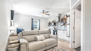 A modern living room with a beige leather sofa, a small side table, and a ceiling fan. A kitchen is visible in the background with white cabinets and stainless steel appliances.