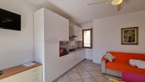 Une cuisine compacte avec des meubles blancs, un four encastré et une fenêtre. On y trouve un petit coin repas avec une table et des chaises, ainsi qu’un canapé agrémenté d’un coussin orange.