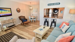 A living room with a sofa, armchairs, a coffee table, a TV, a dining table with chairs, a kitchen area, and a sign that reads "BEACH HOUSE".