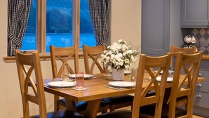 A dining area with a wooden table and chairs, a vase with flowers, and a view of a blue outdoor area through a window.