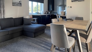 A modern living room with a dark-colored sofa, a dining table with chairs, and a kitchen area with dark cabinets and a window. - Enviable location within the golf course. (Skipton)