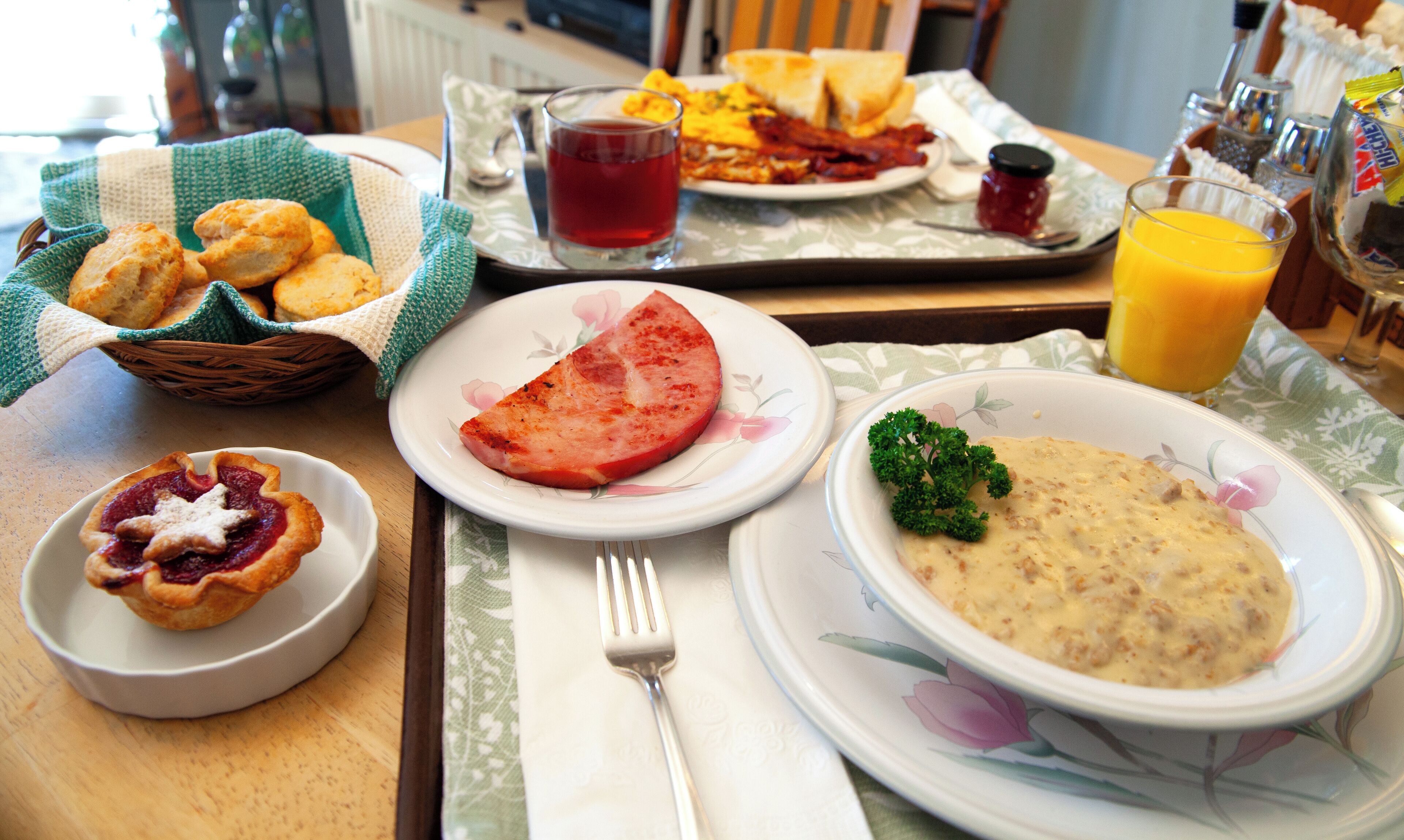 Ett frukostbord med tallrikar fyllda med mat, en korg med bröd, ett glas juice och en kanna med apelsinjuice.