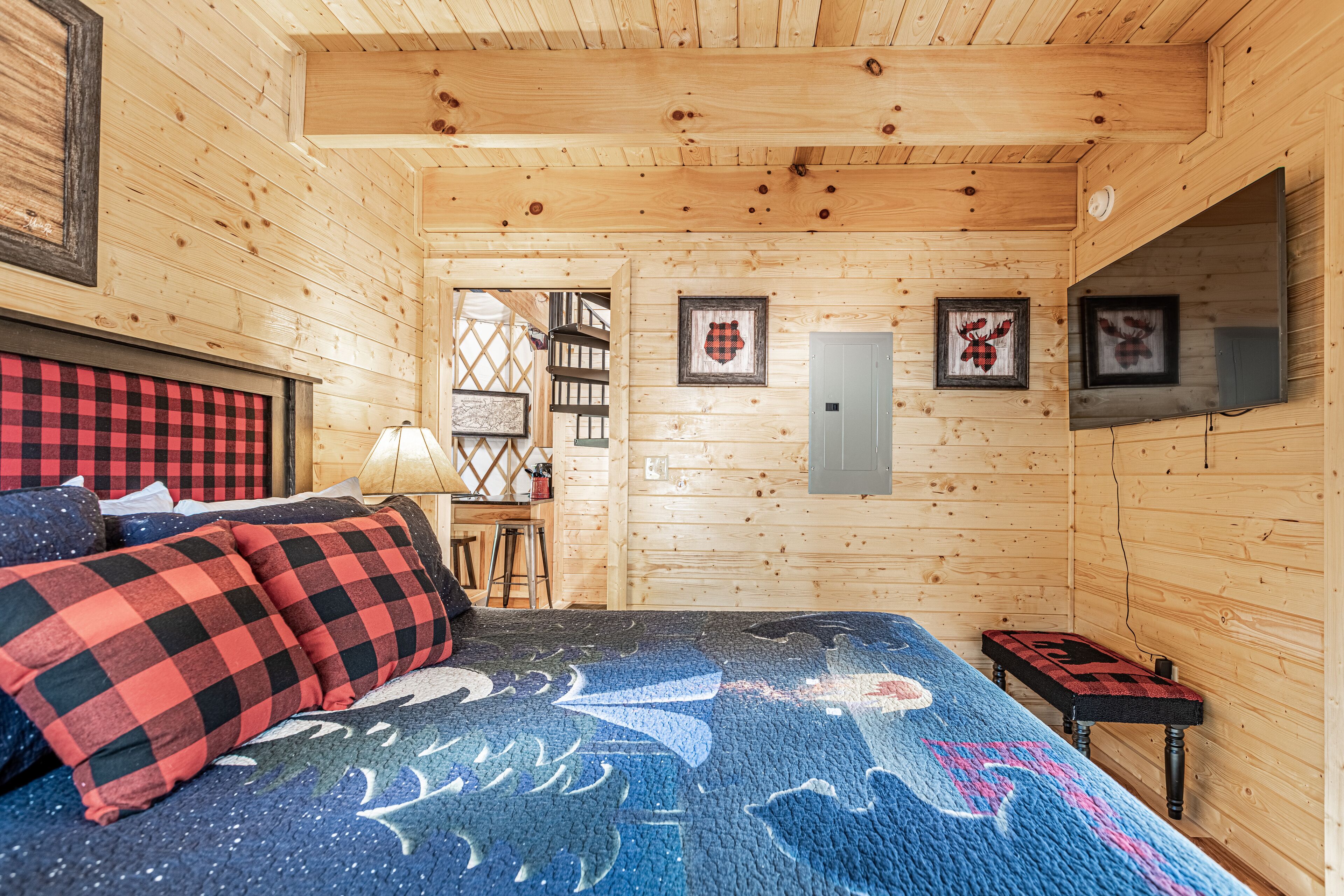 Ein Schlafzimmer einer Holzhütte mit einem Bett, karierten Kissen, einem an der Wand montierten Fernseher und gerahmten Bildern.